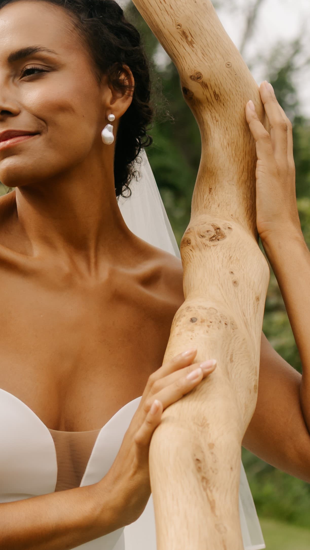 Smiling bride in a white dress and pearl earrings holding a smooth wooden branch.