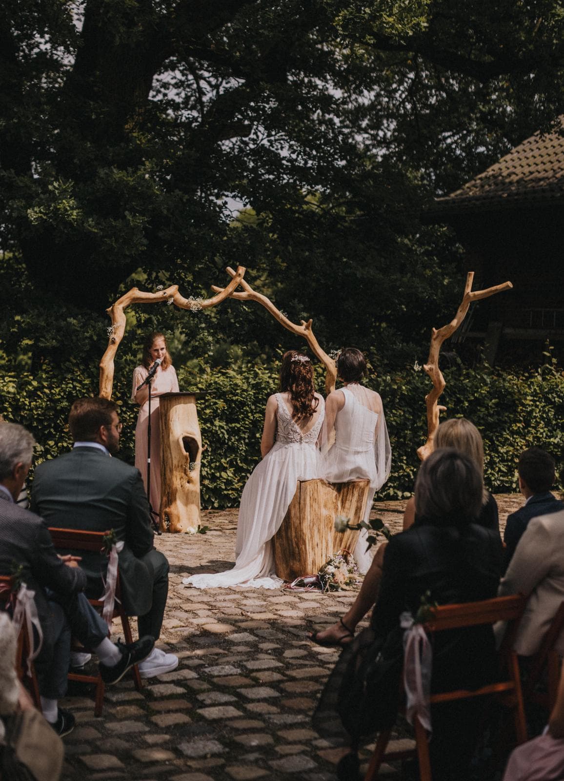 Two brides sit on a tree stump during a rustic outdoor wedding ceremony under branches.