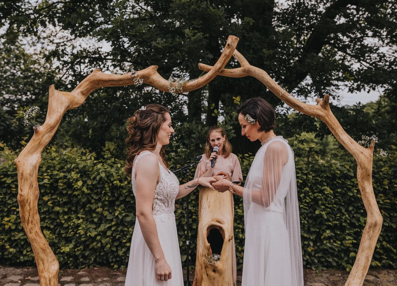 Two brides exchange rings under a rustic wooden arch during an outdoor wedding ceremony.